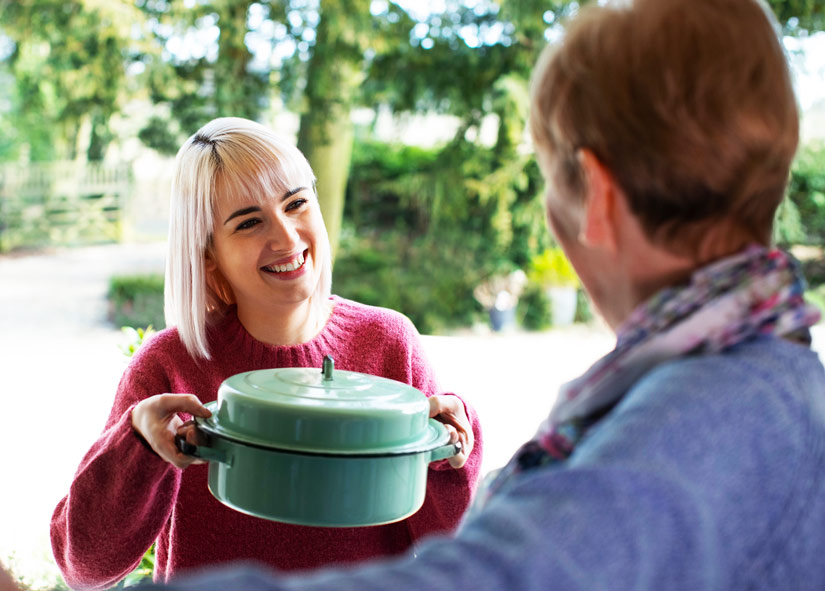 young blonde woman bringing casserole to elderly neighbor at front door - support