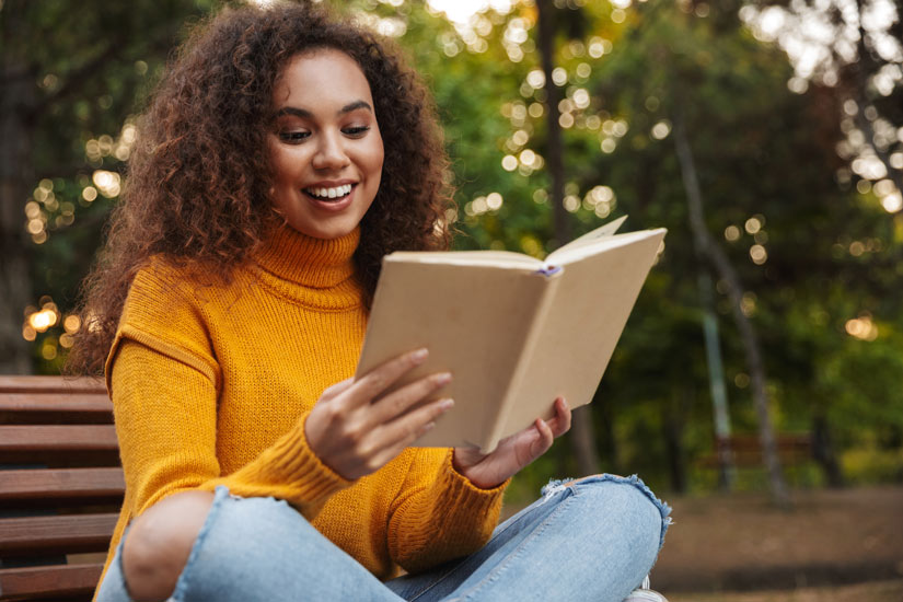beautiful young Black woman in a bright yellow sweater smiling and reading a book outdoors - boredom