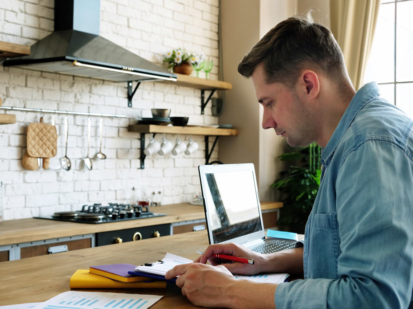 man in his early thirties sitting at home with laptop and files of paperwork - returning to work