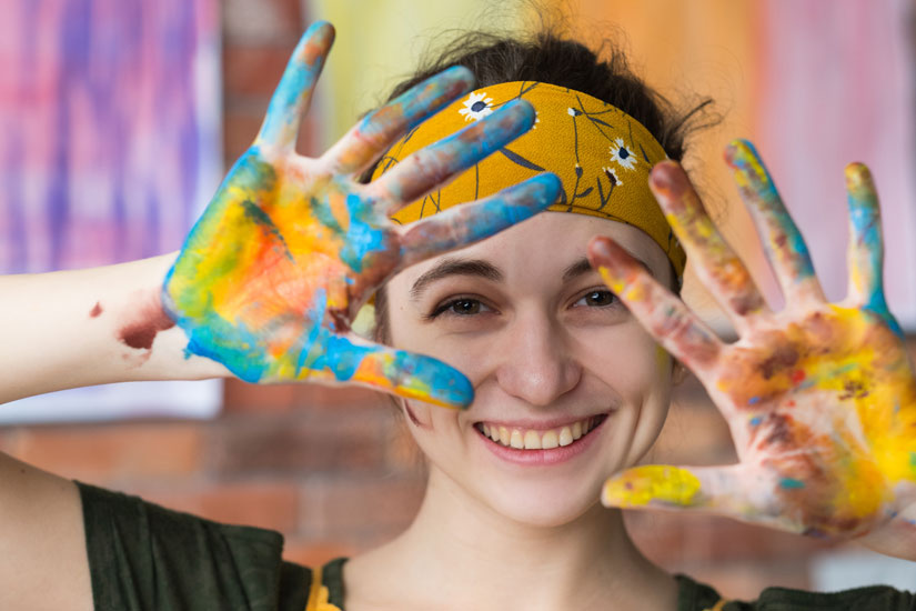 young woman covered in paint, smiling and showing her hands to the camera - experiential art therapy